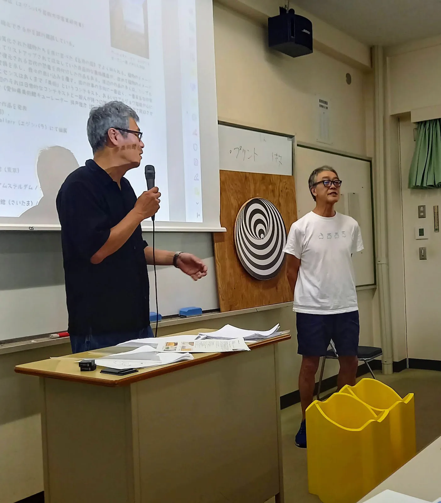 Eiji Watanabe (artist) holding a microphone in a classroom at Aichi University of Education, and Eiji Watanabe (neuroscientist) standing beside an illusion disk